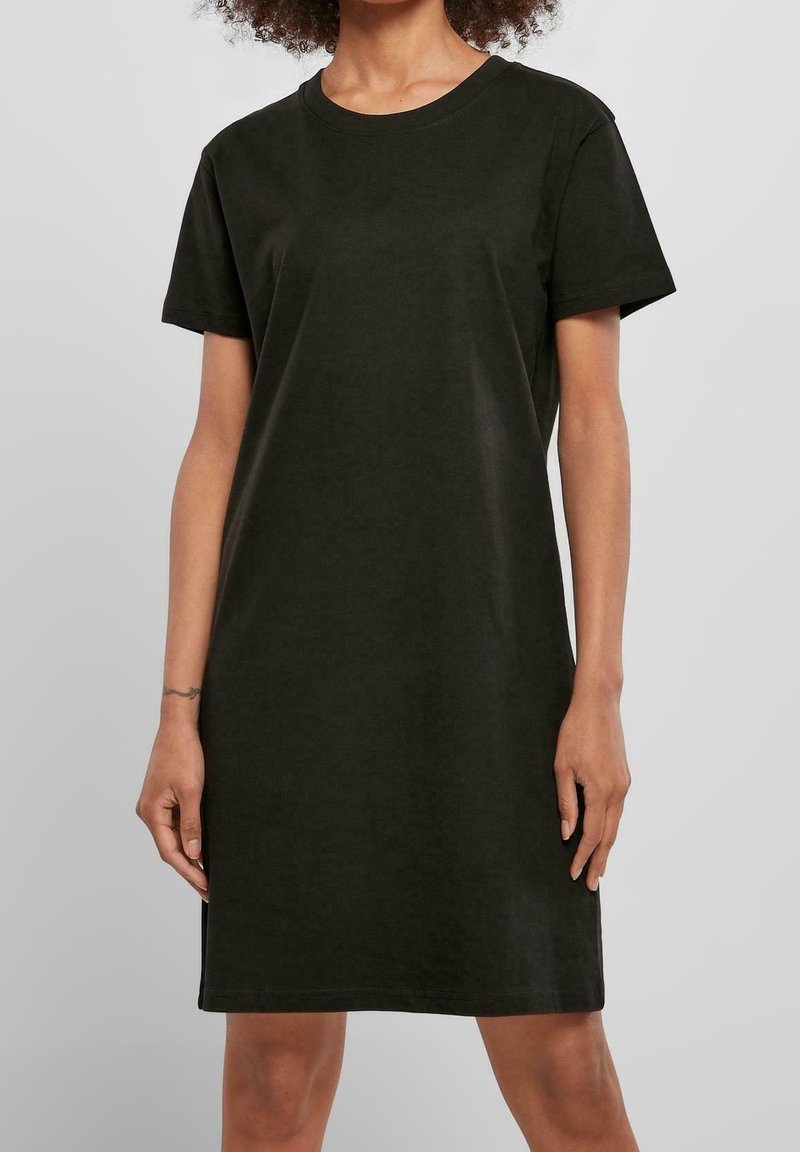 Femme portant une robe T-shirt noire unie à manches courtes et longueur genou, sur un fond neutre.