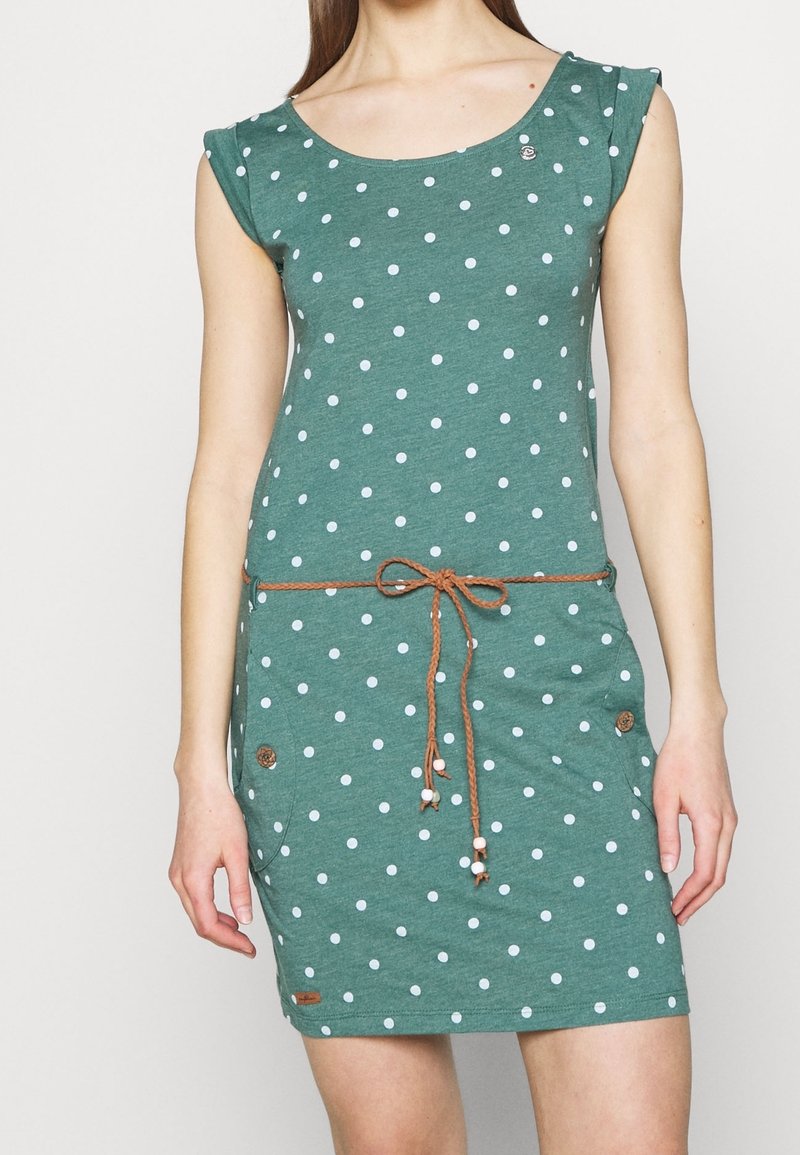 Robe verte à pois blancs, dotée de manches courtes, d'un décolleté en scoop et d'une taille à cordon brun ornée de perles décoratives.