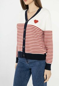 Cardigan rayé rouge et blanc avec bordure bleu marine, col en V, sept boutons et un petit détail en forme de cœur rouge en haut à gauche.