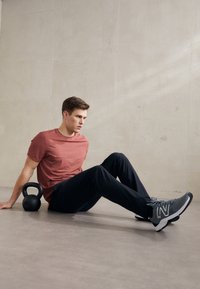 Sporty outfit bestående af en rustfarvet kortærmet skjorte, sorte træningsbukser og grå sneakers med hvide detaljer. Kettlebell ved siden af.