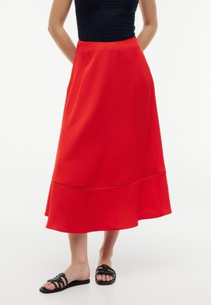 Mujer con falda midi roja de talle alto con un sutil detalle de costura y sandalias planas negras con tiras, de pie sobre un fondo blanco.