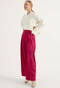Witte lange mouwen, v-hals trui gecombineerd met hoogem waist, geplooide roze broeken. Het model draagt beige hakken en toont een strakke en moderne uitstraling.