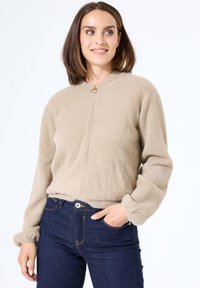 Beige fleece jacket met een ronde halslijn, gouden hardware-accent en elastische manchetten. Gecombineerd met donkerblauwe spijkerbroeken.