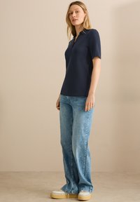 Marineblauwe polo met korte mouwen en een lage V-hals, gecombineerd met lichtblauwe wijde jeans. Glad materiaal, minimalistisch ontwerp en casual stijl.