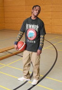 Svart grafisk t-skjorte med teksten "SUMMER LEAGUE", beige bukser og hvite sneakers, holder en basketball, på en gymsal med trebaker.
