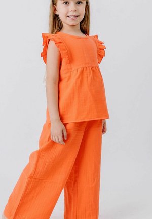 Young girl smiling, wearing bright orange sleeveless top with ruffle details and matching loose pants, standing against plain background.