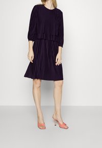 Lila plisserad klänning med trekvartsärmar, lagerdesign; matchad med korallfärgade öppen-tå klackmules med en rosettdetalj.