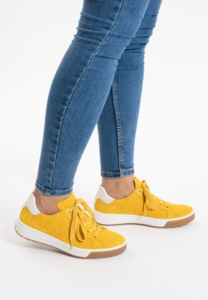 Legs wearing blue skinny jeans and yellow lace-up sneakers with white accents and gum soles against a white background.