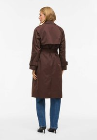 Trench-coat marron avec ceinture, doté d’un grand col et de manches longues, associé à un jean bleu droit et des talons noirs.