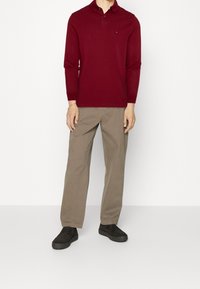 Bordová dlouhá polo košile v kombinaci s pískovými kalhotami a černými teniskami. Košile má límeček a decentní logo na hrudi.