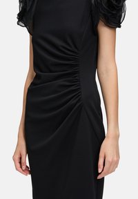 Schwarzes Kleid mit einer figurschmeichelnden Passform, gerafftem Stoff an der Seite und transparenten, gerafften Ärmeln. Glattes Material mit einem leichten Glanz.