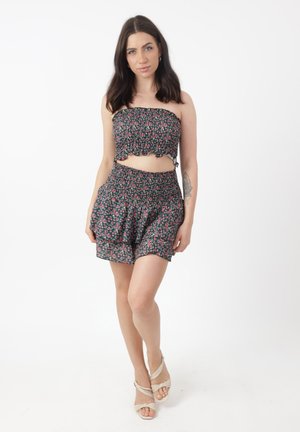 Giovane donna con capelli scuri che indossa un completo coordinato con top a fascia floreale e pantaloncini, in piedi con una gamba incrociata, sandali beige, tatuaggio sul braccio.