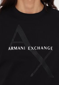 Černé bavlněné tričko s textem "ARMANI EXCHANGE" v bílé barvě, s texturovaným diagonálním grafickým prvkem začleněným do designu.