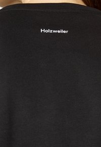 T-shirt noir côtelé avec un logo "Holzweiler" blanc dans le dos. Le matériau est extensible avec une coupe ajustée.