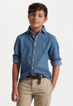 Chemise en denim à boutons avec des manches longues retroussées, couleur bleu clair, arborant un petit logo, associée à un pantalon beige en khaki et une ceinture tressée marron.