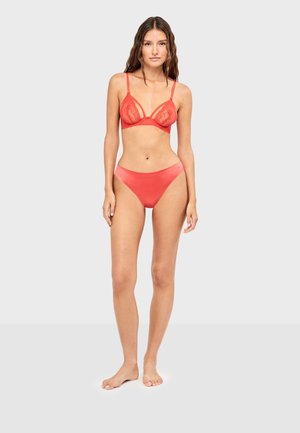 Femme debout pieds nus portant un bralette en dentelle rouge et une culotte en tissu lisse assortie sur un fond clair uni.