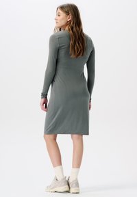 Langärmiges, graues Kleid mit taillierter Passform, knielangem Saum, glatter Textur und minimalen Akzenten. Das Model kombiniert es mit beigen Sneakers und weißen Socken.