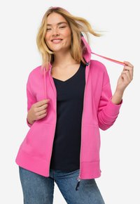 Felpa rosa con zip, tasche a marsupio, cappuccio foderato e stringhe. Abbinata a una maglietta blu navy a V e jeans azzurri chiari.