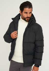 Schwarze Steppjacke mit Kapuze, gesteppter Optik und Reißverschluss vorne. Wird über einem weißen, kabelgestrickten Pullover getragen, mit elastischen Bündchen und Taschen.