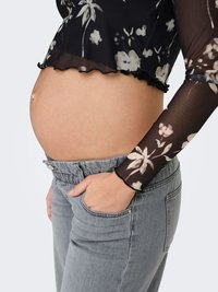 Melna ar ziedu rakstu garroku krekls ar volāniem apakšmalā, kopā ar pelēkām džinsa biksēm ar elastīgu jostasvietu. Redzams grūtnieces puncis, roka atslēgas kabatā.