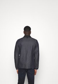 Denim jacket in dark blue with a black collar. Features long sleeves and button cuffs. Clean, minimalist design with no visible patterns.