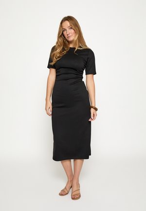 Robe midi noire à manches courtes, coupe ajustée, avec de discrètes fentes latérales. Tissu lisse légèrement brillant, complété par des sandales minimalistes tan.