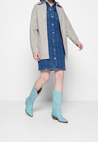 Vestido de mezclilla con cierre de botones en la parte delantera, chaqueta beige de gran tamaño y botas vaqueras azul claro con bordados dorados y diseño de punta afilada.