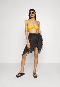 Oransje bikini topp med knute-detalj, sammen med en gjennomsiktig svart sarong med svarte pom-pom aksenter, og kraftige svarte flip flops.