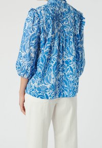 Blouse fleurie bleue avec détails dos froncés, manches trois-quarts bouffantes et tissu léger texturé. Associée à un pantalon clair.