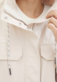 Chaqueta impermeable de color beige claro con cierre frontal, dos bolsillos de parche, capucha ajustable y cordones de contraste con un patrón texturizado.