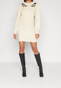 Robe beige en tricot avec des manches longues, broderie florale noire sur les épaules, silhouette cintrée, associée à des bottes noires montantes.