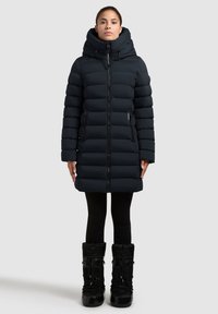 Mujer vestida con un abrigo largo negro de plumas con un alto cuello y capucha, leggings negros y botas de invierno negras, de pie contra un fondo claro.