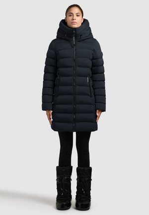 Woman wearing a black long puffer coat with a high collar and hood, black leggings, and black winter boots, standing against a light background.