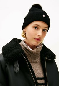 Sort strikket beanie med en pom-pom, som har et lille logo. Laget med en beige ribbet turtleneck under en sort shearling-foret jakke.
