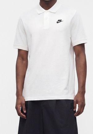 Mężczyzna w białej koszulce polo Nike z czarnym logo po lewej stronie klatki piersiowej i czarnych szortach na jednolitym tle.