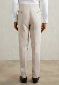 Bézs, szabott nadrág, két gombos hátsó zsebbel, sima textúrájú, egyenes szabású. Fekete cipővel viselve.