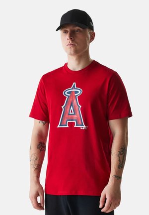 Man draagt een rood Los Angeles Angels T-shirt en een zwarte honkbalpet, staat met een neutrale uitdrukking en zichtbare armtatoeages.