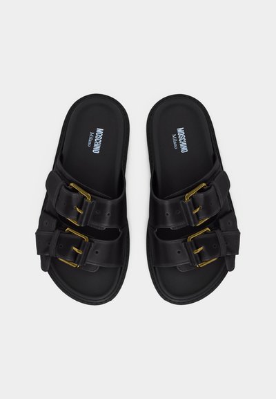 Μαύρα σανδάλια slip-on με διπλές χρυσές αγκράφες, με την επωνυμία "Moschino Milano" στην πατούσα, παρουσιάζονται σε λευκό φόντο.