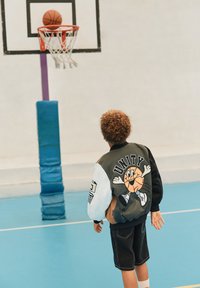 Dreng med krøllet hår iført en "Unity" jakke ser på basketbold, der går gennem kurven på en udendørs blå bane.