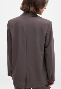Brown tailored blazer with a smooth texture, notched lapels, and three buttons on each cuff. Sleek, structured silhouette.