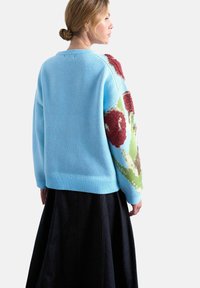 Femme aux cheveux blonds attachés en chignon porte un pull bleu clair avec des motifs floraux rouges et verts sur les manches et une jupe noire.