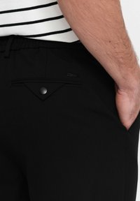 Zwarte broek met een elastische tailleband, beschikt over een achterzak met een driehoekige flap en een zwarte knop. Gladde stof met minimale textuur.
