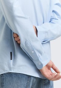 Φωτεινό γαλάζιο πουκάμισο με κουμπιά, μακριά μανίκια, υφή ύφανσης και μανσέτες με κουμπιά. Διαθέτει μικρή ετικέτα στο πλάι. Φορεμένο με τζιν.