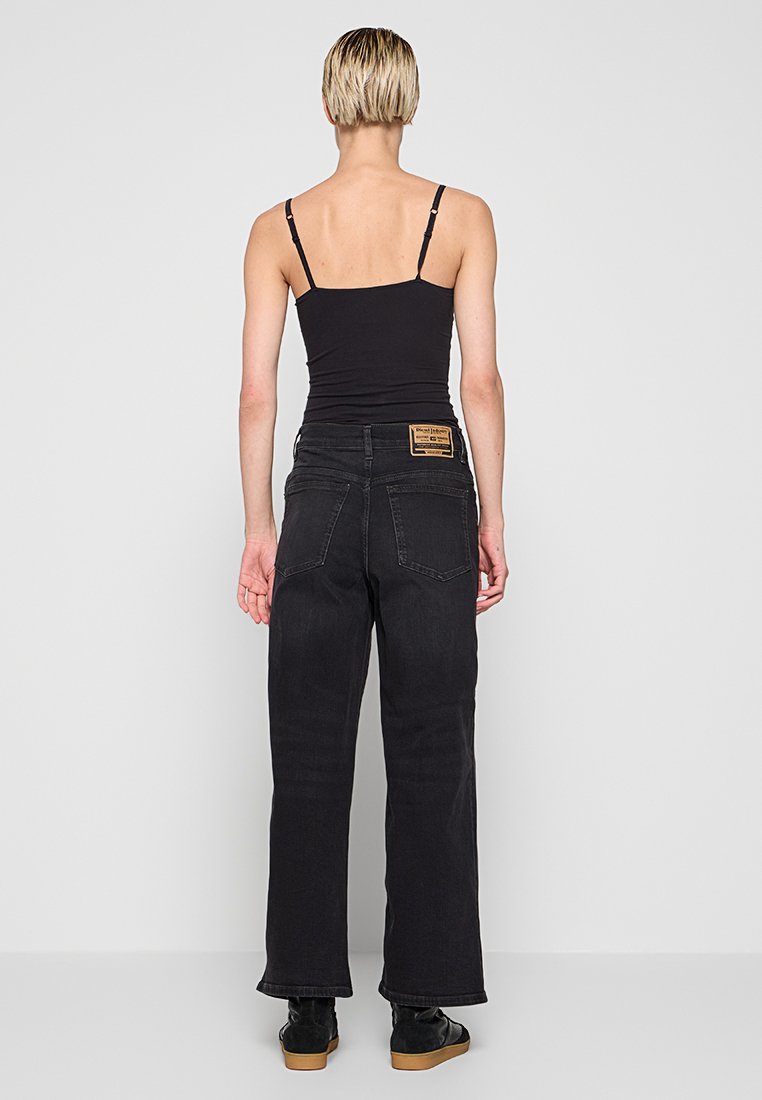 Person mit kurzen blonden Haaren, die ein schwarzes Top mit Spaghettiträgern, hochgeschnittene schwarze Jeans und schwarze Schuhe trägt, mit dem Rücken zur Kamera vor einem grauen Hintergrund.