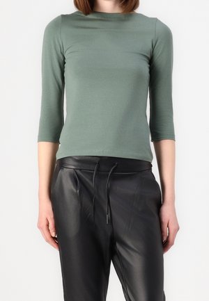 Person iført en grøn tætsiddende langærmet skjorte og sorte læderbukser med høj talje og snøre i livet.