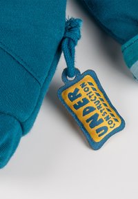 Tessuto blu con un'etichetta cucita gialla e blu recante la scritta "UNDER CONSTRUCTION" attaccata tramite un anello di corda blu.