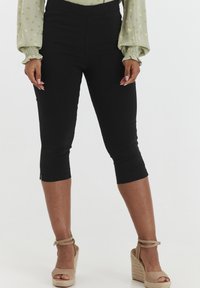 Pantaloni capri neri con una texture liscia, vita elastica e cerniere alla caviglia. Indossati con scarpe espadrilles color sabbia e una blusa floreale leggera.