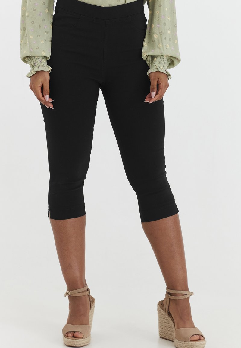 Pantaloni capri neri con una texture liscia, vita elastica e cerniere alla caviglia. Indossati con scarpe espadrilles color sabbia e una blusa floreale leggera.