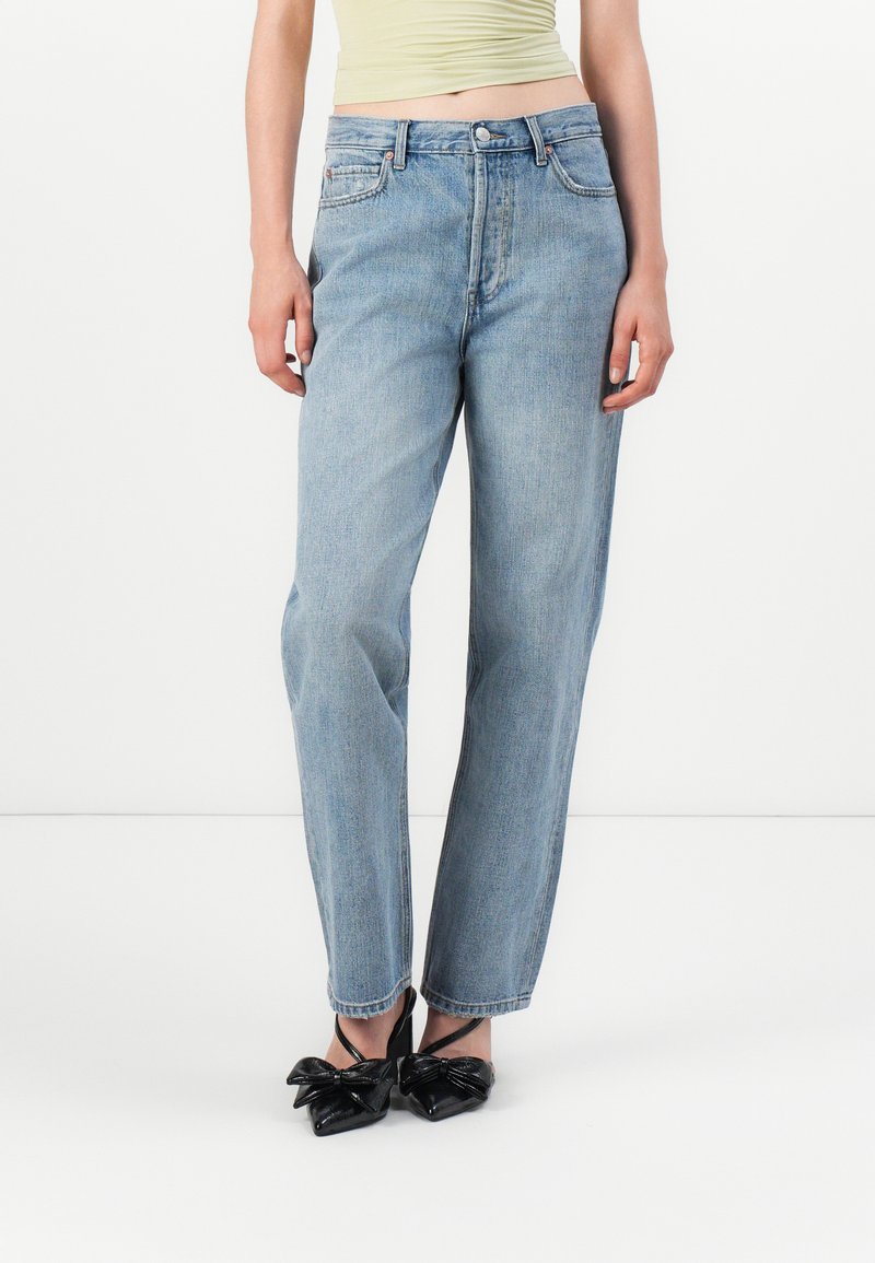 Jeans en denim bleu clair avec une coupe droite, présentant un design classique à cinq poches et un léger délavage. Associés à des chaussures noires à bout pointu.