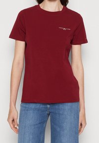 T-shirt in cotone borgogna con scollo rotondo, maniche corte e stampa del logo bianco in alto a sinistra. Abbinata a jeans blu.